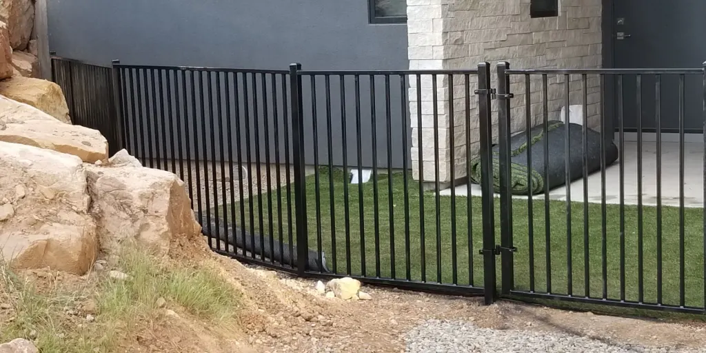 Black ornamental aluminum fence with gate alongside a residential foundation and retaining wall